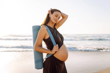 Güneşte sahilde elinde yoga minderiyle gülümseyen hamile kadın. Doğanın tadını çıkaran bir çocuk ve deniz kenarında yürüyüş bekleyen genç bir kadın. Rahatlama, yürüyüş konsepti.
