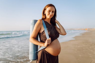 Güneşte sahilde elinde yoga minderiyle gülümseyen hamile kadın. Doğanın tadını çıkaran bir çocuk ve deniz kenarında yürüyüş bekleyen genç bir kadın. Rahatlama, yürüyüş konsepti.