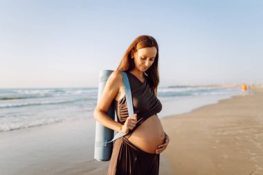 Güneşte sahilde elinde yoga minderiyle gülümseyen hamile kadın. Doğanın tadını çıkaran bir çocuk ve deniz kenarında yürüyüş bekleyen genç bir kadın. Rahatlama, yürüyüş konsepti.