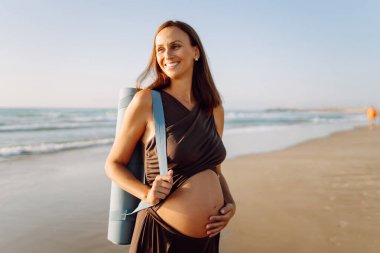 Güneşte sahilde elinde yoga minderiyle gülümseyen hamile kadın. Doğanın tadını çıkaran bir çocuk ve deniz kenarında yürüyüş bekleyen genç bir kadın. Rahatlama, yürüyüş konsepti.
