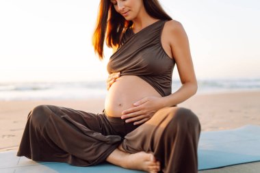 Bir kadın yoga minderinde oturur ve gün batımında hamile karnına dokunur. Deniz kıyısında yoga ve meditasyon yapan genç bir kadının yakın çekimi. Üreme kavramı, sağlık.