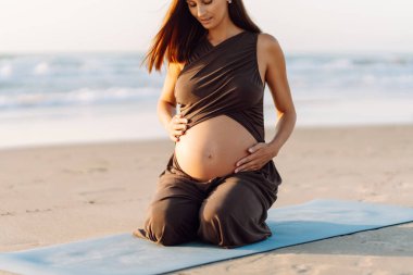Bir kadın yoga minderinde oturur ve gün batımında hamile karnına dokunur. Deniz kıyısında yoga ve meditasyon yapan genç bir kadının yakın çekimi. Üreme kavramı, sağlık.