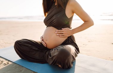 Bir kadın yoga minderinde oturur ve gün batımında hamile karnına dokunur. Deniz kıyısında yoga ve meditasyon yapan genç bir kadının yakın çekimi. Üreme kavramı, sağlık.