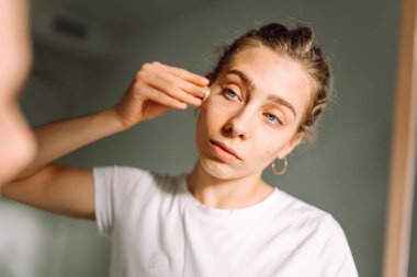 Genç bir kadının yüzünü pamuklu bezle silip aynaya bakarken yaklaş. Hoş bir kadın doğal güzellikten zevk alıyor, rahat bir odada makyaj yapıyor. Güzellik, cilt bakımı.