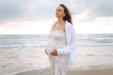 Elbise giymiş hamile bir kadın deniz kıyısında yürüyor, karnına dokunuyor. Güzel bir kadın doğadan zevk alır, özgürlüğü hisseder. Yürüyüş konsepti, hafta sonu..