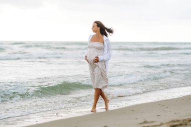 Elbise giymiş hamile bir kadın deniz kıyısında yürüyor, karnına dokunuyor. Güzel bir kadın doğadan zevk alır, özgürlüğü hisseder. Yürüyüş konsepti, hafta sonu..