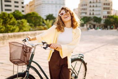 Güneşli bir şehirde bisikletli mutlu bir kadın. Güneş gözlüklü turist bisiklet sürmeyi, şehir atmosferini seviyor. Blogculuk, turizm konsepti. Etkin yaşam biçimi.