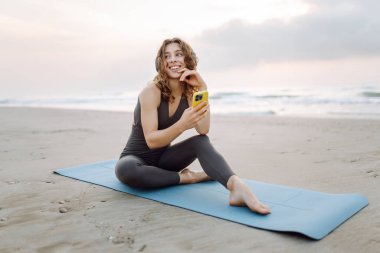 Kumsalda yoga minderinde oturan telefonlu ve kablosuz kulaklıklı, spor kıyafetli gülümseyen kadın. Egzersiz sonrası müzik ve deniz manzarası dinlemekten hoşlanan genç bir kadın. Doğa, teknoloji konsepti.