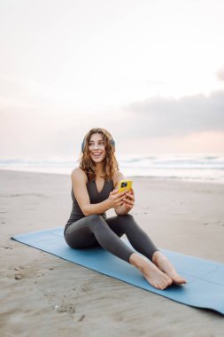Kumsalda yoga minderinde oturan telefonlu ve kablosuz kulaklıklı, spor kıyafetli gülümseyen kadın. Egzersiz sonrası müzik ve deniz manzarası dinlemekten hoşlanan genç bir kadın. Doğa, teknoloji konsepti.