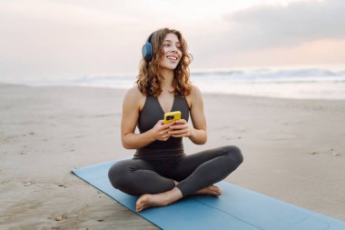 Kumsalda yoga minderinde oturan telefonlu ve kablosuz kulaklıklı, spor kıyafetli gülümseyen kadın. Egzersiz sonrası müzik ve deniz manzarası dinlemekten hoşlanan genç bir kadın. Doğa, teknoloji konsepti.