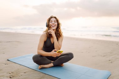 Kumsalda yoga minderinde oturan telefonlu ve kablosuz kulaklıklı, spor kıyafetli gülümseyen kadın. Egzersiz sonrası müzik ve deniz manzarası dinlemekten hoşlanan genç bir kadın. Doğa, teknoloji konsepti.