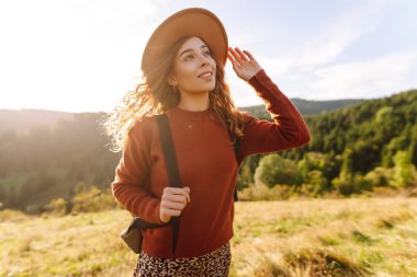 Sırt çantası ve şapkası olan neşeli bir kadın gün batımında yeşil tepelerde yürüyor. Bir gezgin dağlarda doğanın tadını çıkarır ve özgür hisseder. Etkin yaşam biçimi.