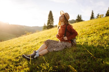 Sırt çantası ve şapkası olan neşeli bir kadın gün batımında yeşil tepelerde yürüyor. Bir gezgin dağlarda doğanın tadını çıkarır ve özgür hisseder. Etkin yaşam biçimi.