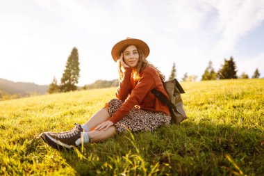Sırt çantası ve şapkası olan neşeli bir kadın gün batımında yeşil tepelerde yürüyor. Bir gezgin dağlarda doğanın tadını çıkarır ve özgür hisseder. Etkin yaşam biçimi.