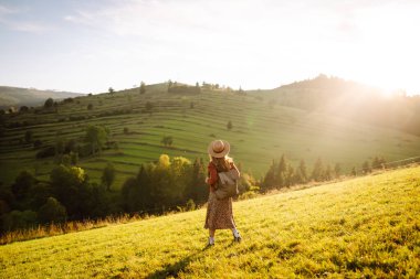 Sırt çantası ve şapkası olan neşeli bir kadın gün batımında yeşil tepelerde yürüyor. Bir gezgin dağlarda doğanın tadını çıkarır ve özgür hisseder. Etkin yaşam biçimi.