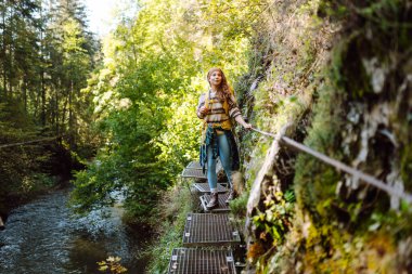 Sırt çantalı kadın yürüyüşçü dağlardaki yaya asma köprüsünde yürüyor. Genç bir kadın dağ manzarasından zevk alıyor ve açık havada özgürlüğü hissediyor. Seyahat, özgürlük kavramı.
