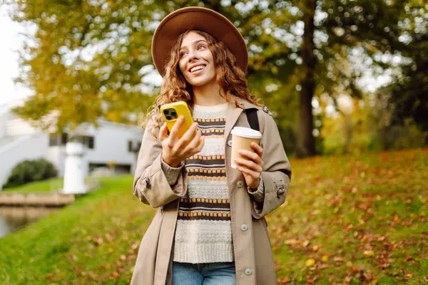 Bir sonbahar parkında yürürken elinde bir fincan sıcak içecek ve telefon tutan şapkalı genç bir kadının portresi. Neşeli turist blogları, kahve içmek ve güneşli bir günün tadını çıkarmak. Yaşam biçimi.