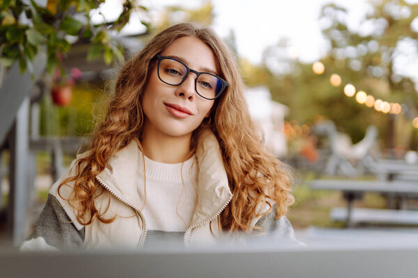 Портрет молодой женщины в очках, сидящей за деревянным столом с ноутбуком в осеннем парке. Хорошая женщина-фрилансер, наслаждающаяся свежим воздухом и работающая с ноутбуком. Концепция удаленной работы, выходные.