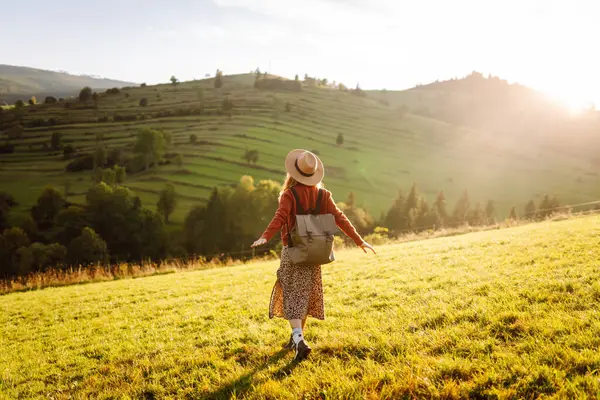 Sırt çantası ve şapkası olan neşeli bir kadın gün batımında yeşil tepelerde yürüyor. Bir gezgin dağlarda doğanın tadını çıkarır ve özgür hisseder. Etkin yaşam biçimi.