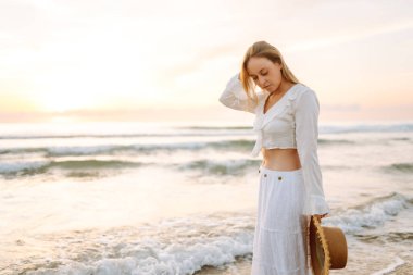 A young woman strolls along a sandy beach at sunrise. A beautiful woman in a hat enjoys the seascape, feeling free. Concept: vacation, weekend, and enjoyment.