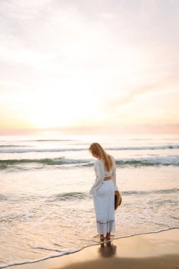 A young woman strolls along a sandy beach at sunrise. A beautiful woman in a hat enjoys the seascape, feeling free. Concept: vacation, weekend, and enjoyment.