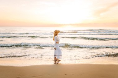 A young woman strolls along a sandy beach at sunrise. A beautiful woman in a hat enjoys the seascape, feeling free. Concept: vacation, weekend, and enjoyment.