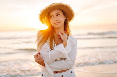 A young woman strolls along a sandy beach at sunrise. A beautiful woman in a hat enjoys the seascape, feeling free. Concept: vacation, weekend, and enjoyment.