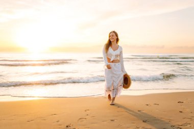 A young woman strolls along a sandy beach at sunrise. A beautiful woman in a hat enjoys the seascape, feeling free. Concept: vacation, weekend, and enjoyment.