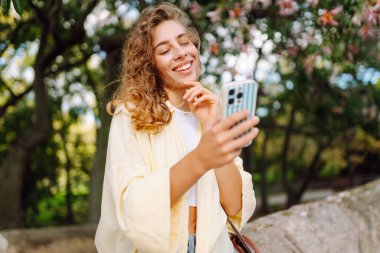 A cheerful woman with a phone takes a stroll on a sunny day. A beautiful woman enjoys the weather, writing a blog or sending messages outdoors. Concept of walking, relaxation, and technology.