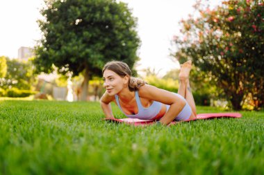A young woman in activewear exercises in a sunny park. An athletic woman performs exercises on a pink yoga mat on a green lawn. Concepts: sports, outdoors, active lifestyle.