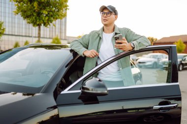 A young man wearing glasses and holding a phone stands next to a car in a parking lot. The man enjoys the sunny day with his phone next to a dark car. Conceptual image: transportation, leisure.