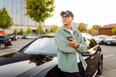 A young man wearing glasses and holding a phone stands next to a car in a parking lot. The man enjoys the sunny day with his phone next to a dark car. Conceptual image: transportation, leisure.