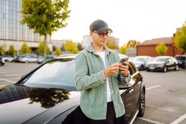 A young man wearing glasses and holding a phone stands next to a car in a parking lot. The man enjoys the sunny day with his phone next to a dark car. Conceptual image: transportation, leisure.
