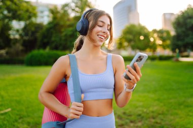 A smiling woman in athletic clothes stands in a park with a bright yoga mat. An athletic woman after a workout listens to music with her phone in the sun's rays. Concept of enjoyment and health.