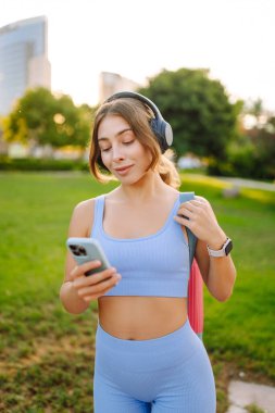 A smiling woman in athletic clothes stands in a park with a bright yoga mat. An athletic woman after a workout listens to music with her phone in the sun's rays. Concept of enjoyment and health.