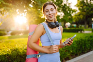 A smiling woman in athletic clothes stands in a park with a bright yoga mat. An athletic woman after a workout listens to music with her phone in the sun's rays. Concept of enjoyment and health.