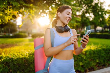A smiling woman in athletic clothes stands in a park with a bright yoga mat. An athletic woman after a workout listens to music with her phone in the sun's rays. Concept of enjoyment and health.