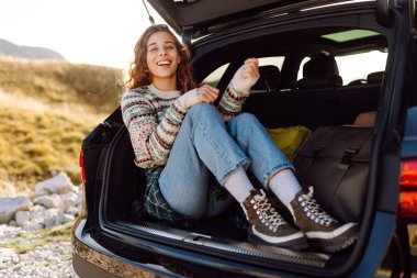 A young traveler sits in the trunk of a car on a sunny day. A beautiful woman enjoys the mountain scenery on a road trip. Concept of adventure, nature, and relaxation.