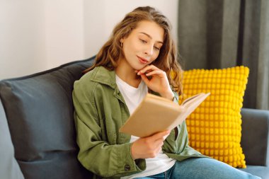 A young woman reads a book while sitting on the floor in a cozy room. A beautiful woman studies in the living room near the sofa, enjoying the space. Concepts of education, learning, and comfort.