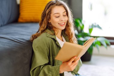 A young woman reads a book while sitting on the floor in a cozy room. A beautiful woman studies in the living room near the sofa, enjoying the space. Concepts of education, learning, and comfort.