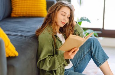 A young woman reads a book while sitting on the floor in a cozy room. A beautiful woman studies in the living room near the sofa, enjoying the space. Concepts of education, learning, and comfort.
