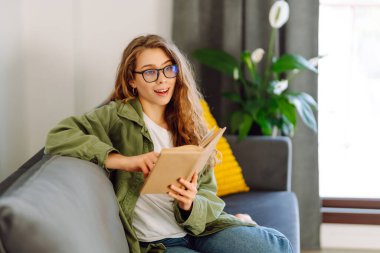 A young woman reads a book while sitting on the floor in a cozy room. A beautiful woman studies in the living room near the sofa, enjoying the space. Concepts of education, learning, and comfort.