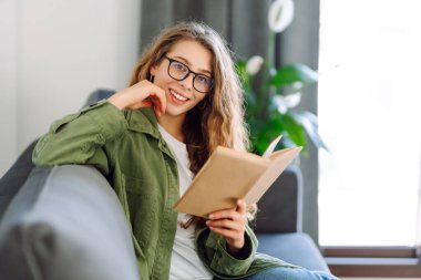 A young woman reads a book while sitting on the floor in a cozy room. A beautiful woman studies in the living room near the sofa, enjoying the space. Concepts of education, learning, and comfort.