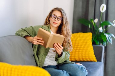 A young woman reads a book while sitting on the floor in a cozy room. A beautiful woman studies in the living room near the sofa, enjoying the space. Concepts of education, learning, and comfort.