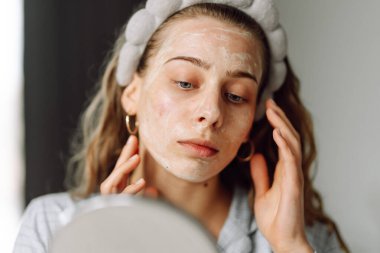 A close-up of a young woman's face with a mask reflected in a small mirror. A beautiful woman pampers herself in a cozy room. Concepts: skincare, beauty, self-care.