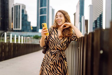 A young woman with phone enjoys life against the backdrop of urban metropolis. Beautiful tourist strolls along pedestrian bridge with phone, texting or blogging. Concept of the city, technology.