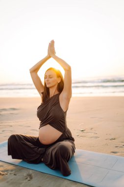 A pregnant woman meditates on beach in sun's rays. A beautiful expectant mother performs yoga on yoga mat at sunrise. Concepts of health, motherhood, an active lifestyle.