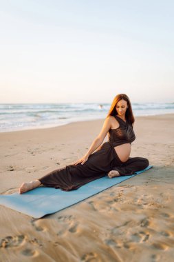 A pregnant woman meditates on beach in sun's rays. A beautiful expectant mother performs yoga on yoga mat at sunrise. Concepts of health, motherhood, an active lifestyle.