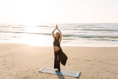 A pregnant woman meditates on beach in sun's rays. A beautiful expectant mother performs yoga on yoga mat at sunrise. Concepts of health, motherhood, an active lifestyle.