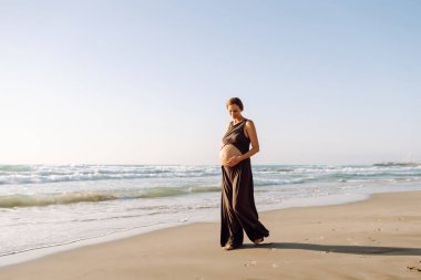 A pregnant woman meditates on beach in sun's rays. A beautiful expectant mother performs yoga on yoga mat at sunrise. Concepts of health, motherhood, an active lifestyle.